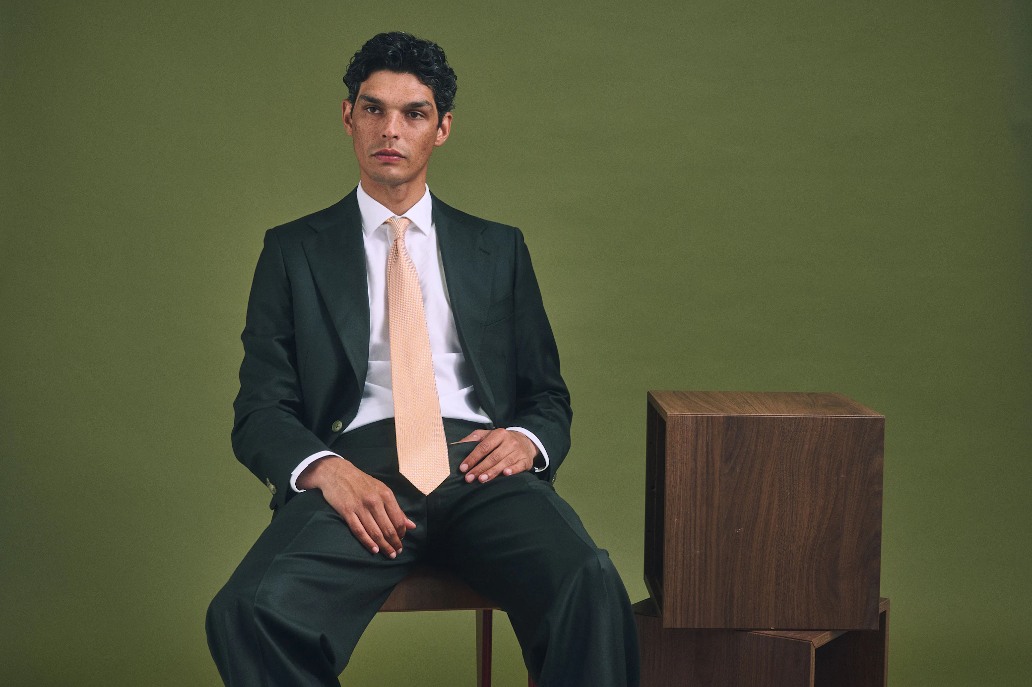 The Alagna Loro Piana forest green wool suit worn seated beside a timber plinth