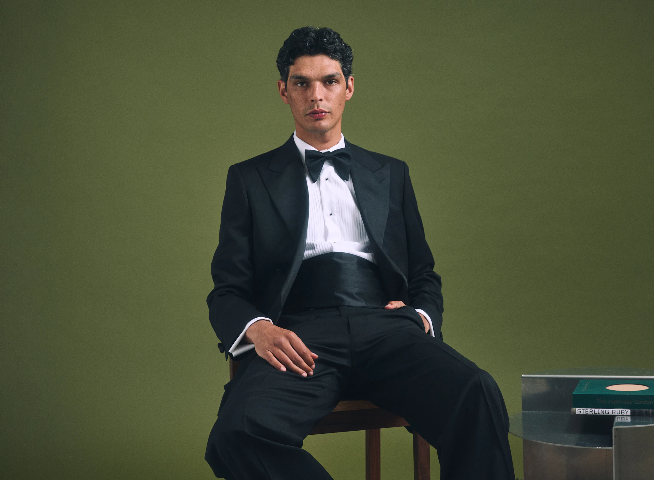 A man wearing the Quarona Loro Piana black wool tuxedo, seated against an olive backdrop