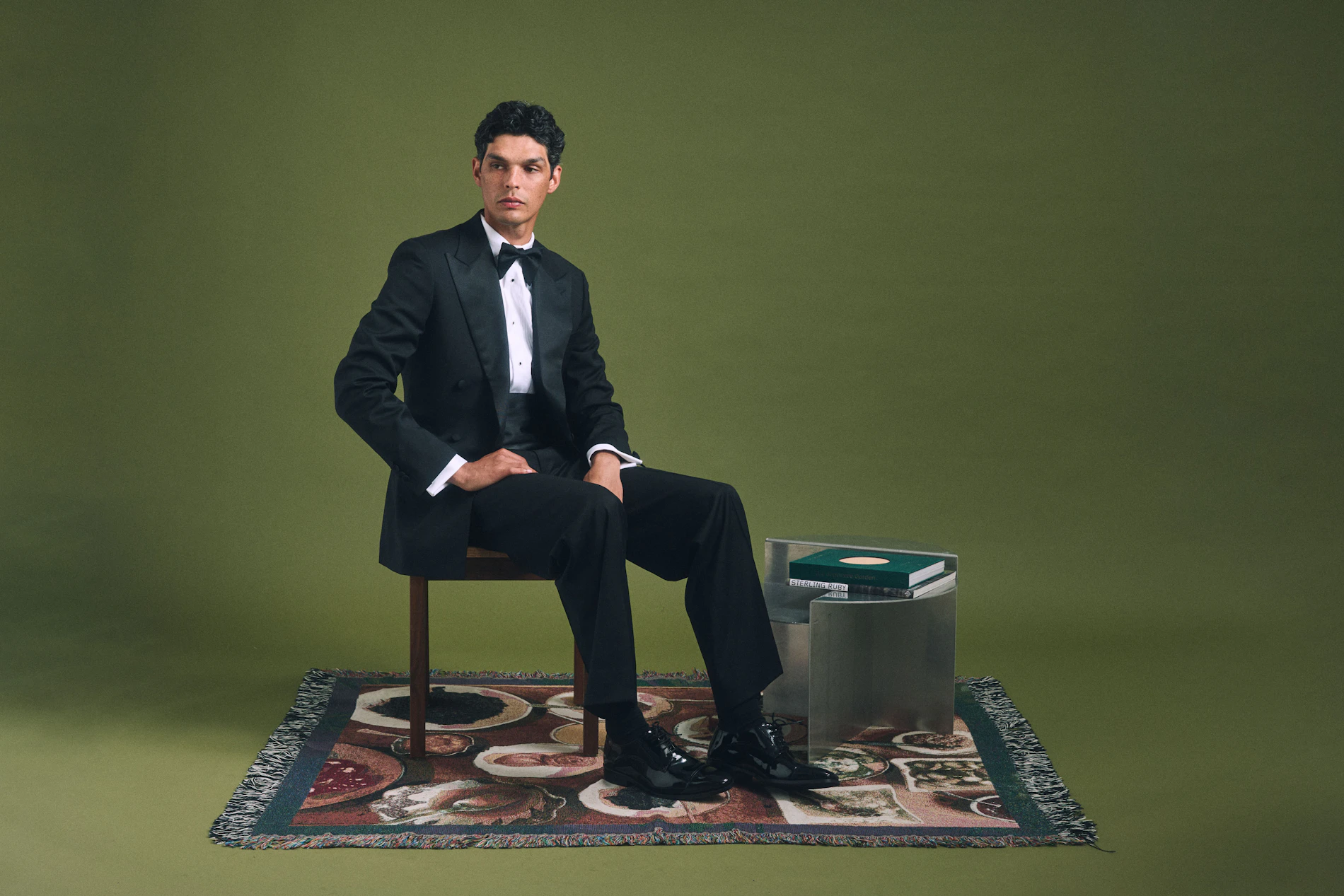 The Quarona Loro Piana black wool tuxedo worn seated on a patterned rug