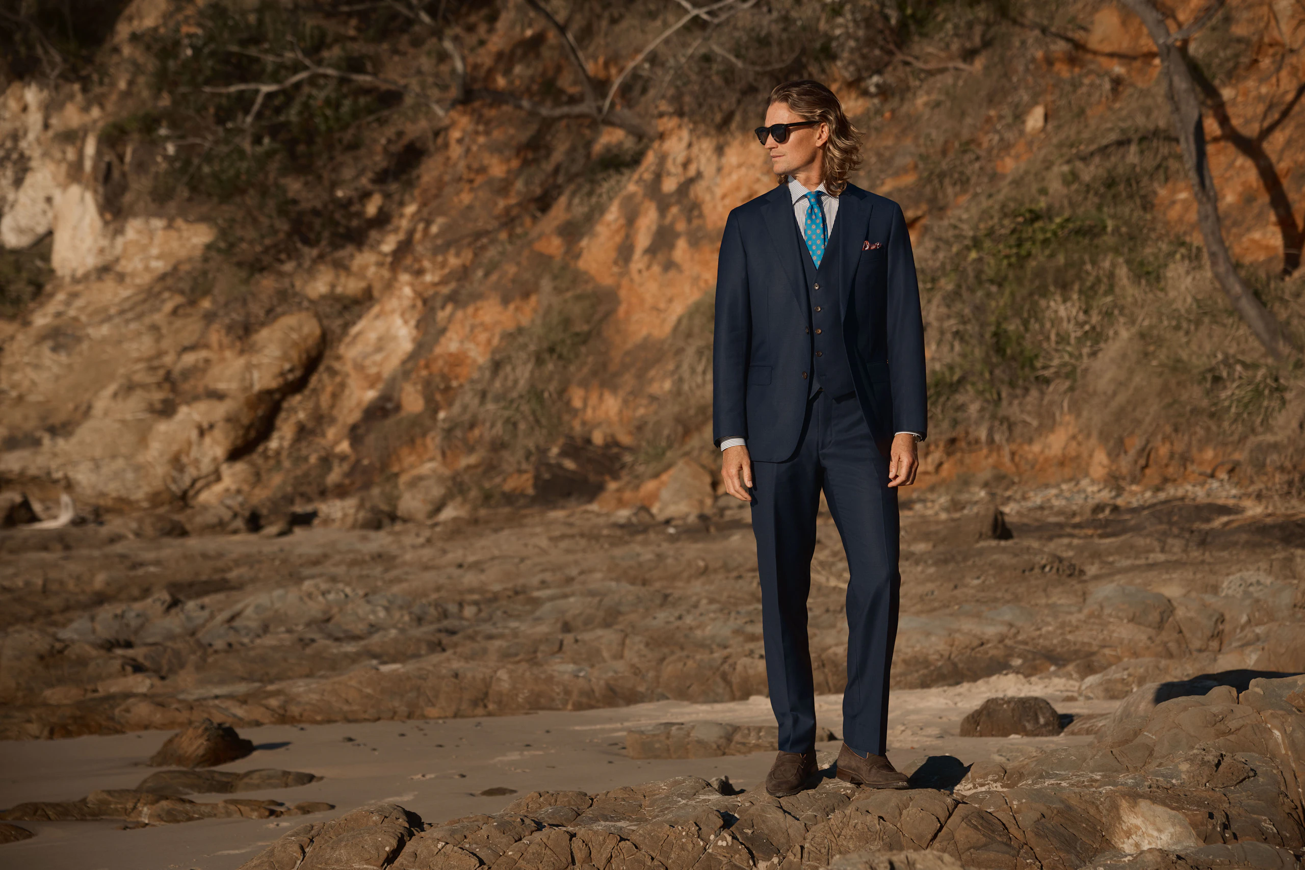 A man in a navy three-piece suit standing on a rocky beach
