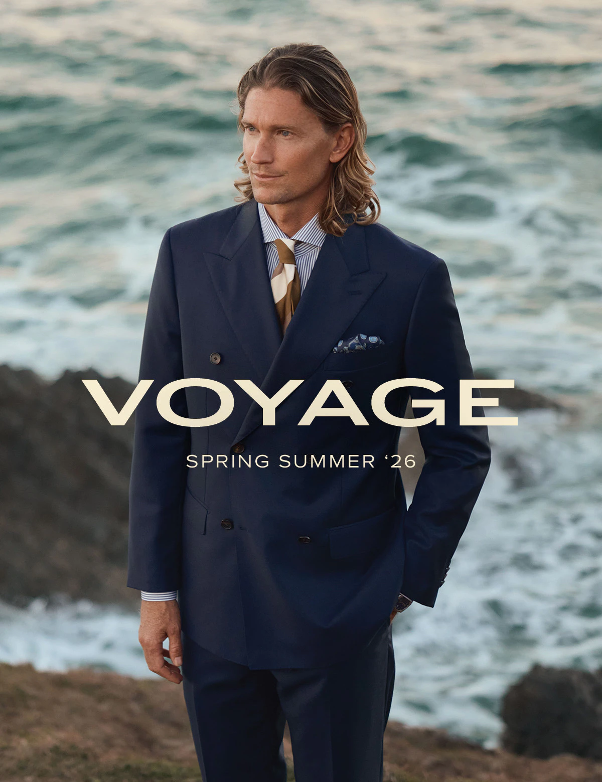 A man in a navy double-breasted suit standing before the surf, with the Voyage Spring Summer '26 title over the water