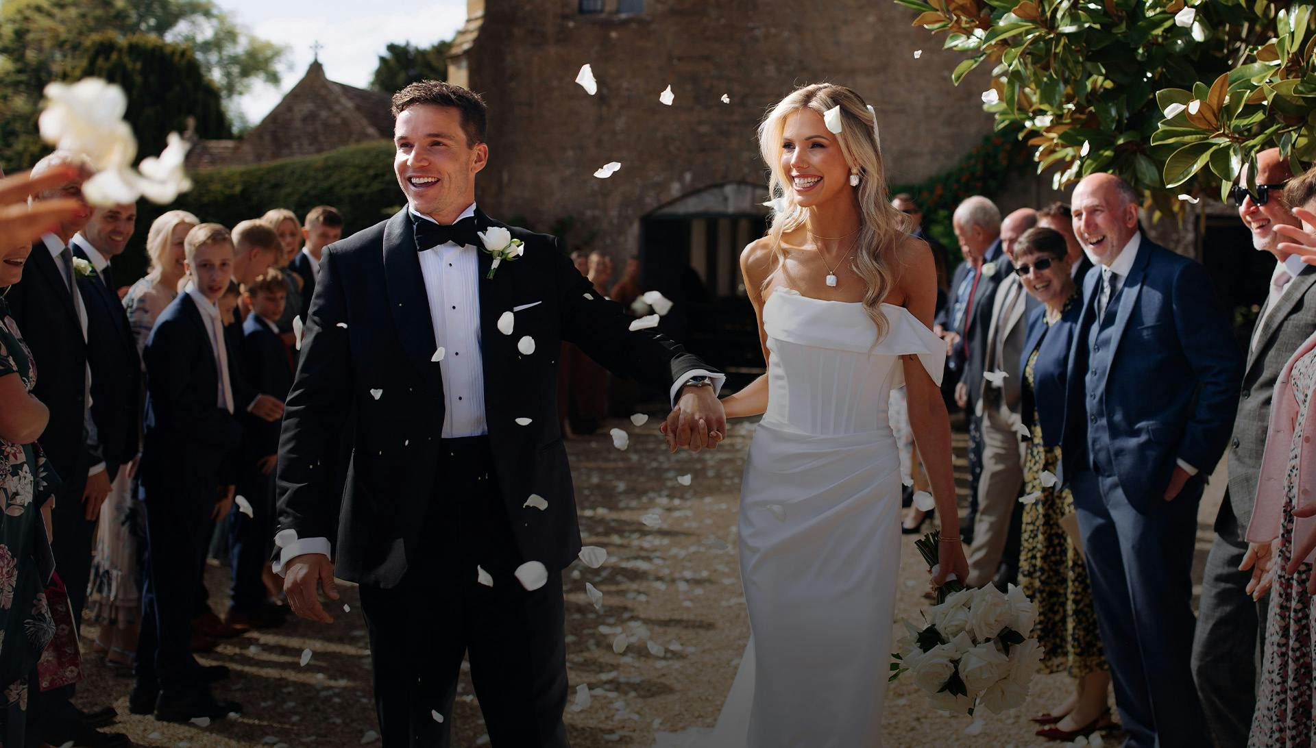 Bride and groom holding hands while guests throw white rose petals, the groom wearing a custom black tuxedo
